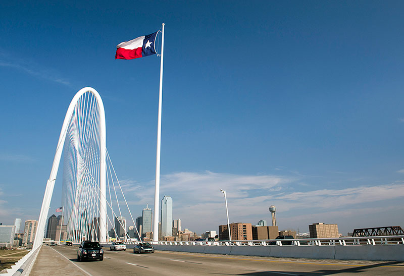 The Margaret Hunt Hill Bridge in Dallas, TX with the Texas flag flowing in front of the skyline.